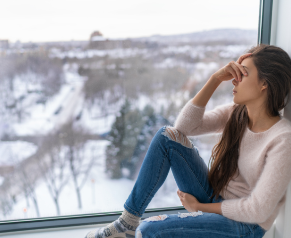 Vrouw die verdrietig bij een raam zit en haar gezicht vasthoudt, als weergave van emotionele impact van Seksuele disfuncties en intimiteitsproblemen en het ervaren van angst, onzekerheid en mentale spanning.